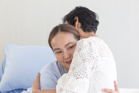 A young Asian woman who is sick in a hospital embraces her mother who looks after her.