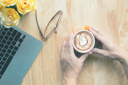 Hand hold Cup of coffee with beautiful Latte art,laptop,flower on work table,top view vintage tone.の写真素材