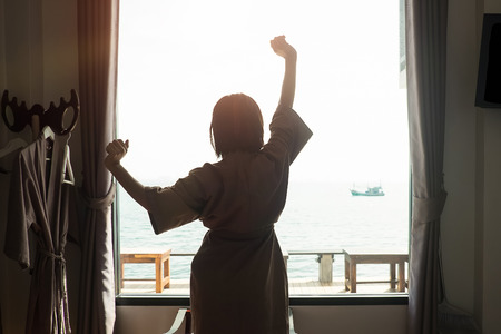 Holiday concept Silhouette photo of woman posting relax action near a windows and outside is a sea with vintage and dark tone, soft focus photo.の写真素材