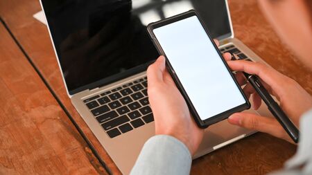 Cropped shot of businessman holding a pen and white blank screen smartphone in front laptop with the wooden working desk as background.の写真素材