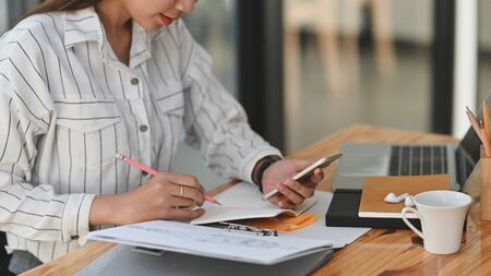 Cropped shot secretary woman in white striped shirt sitting at the wooden desk while writing on the...