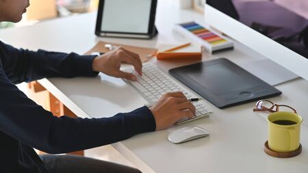 Side shot of young creative man while typing on the keyboard. Including Coffee cup, mouse, keyboard, color guide, tablet earphone, notebook and pencil on the work desk.の写真素材