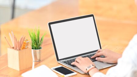 Cropped shot of young creative woman surfing the internet by using white blank screen laptop while sitting at the modern wooden desk.の写真素材