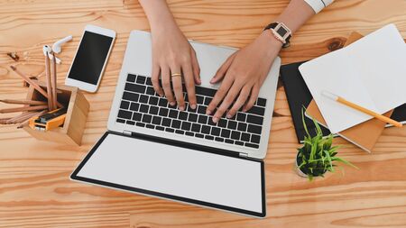 Top view of woman hand while using/typing on black blank screen laptop including pencil holder, black blank screen smartphone, potted plant, pencil, notes, earphone on wooden desk.の写真素材