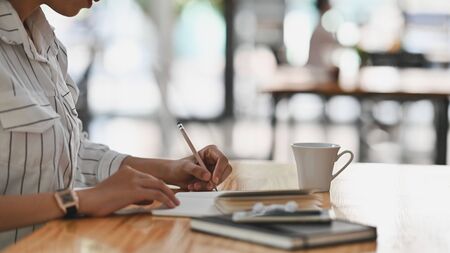 Cropped shot of young secretary woman in white striped shirt sitting at the wooden desk and writing...