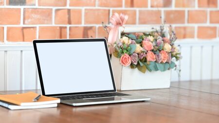 Photo of white blank screen laptop, notebook, pen and roses bouquet in the white plastic basket putting on the wooden table with modern concrete wall as background.の写真素材