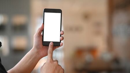 Man's hand holding crop black smartphone with white blank screen and pointing finger on button with modern living room as blurred background.の写真素材