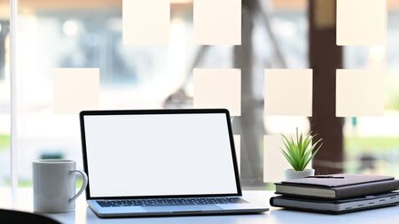 Laptop with white blank screen, potted plant, coffee cup and stack of books putting together on modern working desk with blurred background.の写真素材