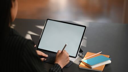 Cropped shot through shoulder of business woman typing on keyboard of tablet and holding electric pen in hand while sitting at the modern working table with the comfortable office room as background.の写真素材