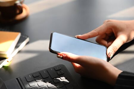 Cropped shot of businesswoman's hand while holding smartphone with white blank screen on the modern working desk close to the tablet with keyboard case, notebook and coffee cup.の写真素材