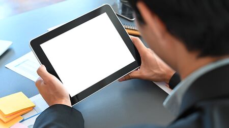 Cropped shot of smart businessman in black suit holding crop black tablet with white blank screen in hand while sitting at the modern table with modern meeting room as background.の写真素材