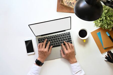 Top view of creative man typing on white blank screen laptop on the modern working desk. Smartphone with black empty display, coffee cup, notebook, lamp, pencil holder and potted plant putting on it.の写真素材