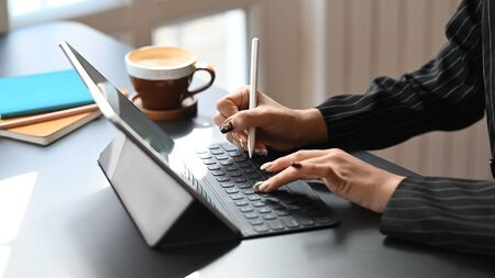 Cropped shot of business woman typing on keyboard of tablet and holding electric pen in hand while sitting at the modern working table with the comfortable office room as background.の写真素材