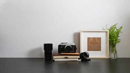 Photographer workspace with white wall as background. Camera, Lens, Stack of books, Picture frame and Potted plant putting together on black wooden table in orderly living room.の写真素材
