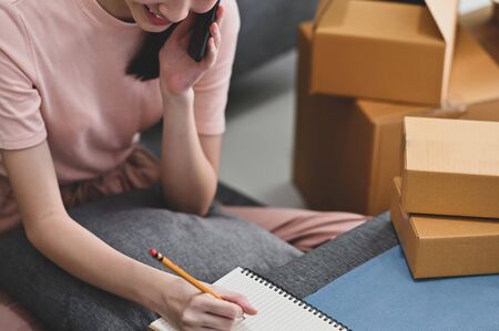 Cropped shot young beautiful girl talking on her smartphone and writing on notebook while sitting at...