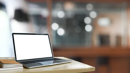 Photo of Computer laptop with white blank screen and books putting together on modern wooden table with blurred living room as background. Comfortable/Orderly work space concept. Space for advertise.の写真素材