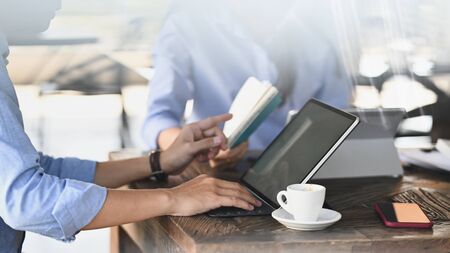 Cropped image of smart man working as secretary typing on computer tablet and pointing his finger to notebook while sitting together with his colleague at the modern wooden table.の写真素材