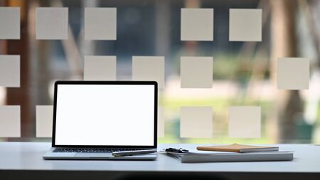 Photo of Computer laptop with white blank screen, notebook, clipboard and pen putting together on white modern desk with blurred wall glass as background. Space for advertise concept.の写真素材
