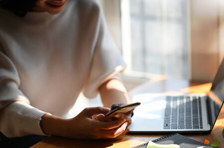 Cropped shot of young designer woman holding smartphone in hands while sitting at the modern wooden...
