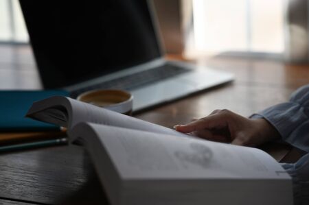 Closeup woman sitting at the modern wooden table with the coffee cup and reading a book in her hand. Relaxing time, Morning vibe concept. Female lifestyle concept.の写真素材
