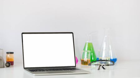 Laboratory glassware containing colored liquids, Computer laptop with white isolate screen and safety glasses while putting together on white table isolated over white background.の写真素材