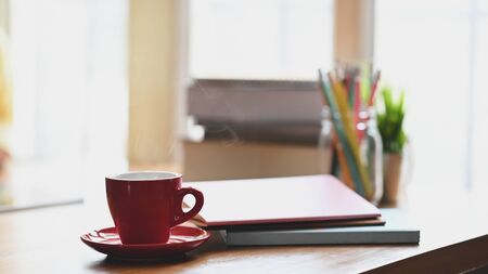 Coffee morning concept, Red coffee cup with coasters putting on wooden table with pencil holder, stack of books, potted plant over living room windows as background.の写真素材