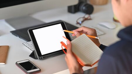 Cropped shot behind of smart man take notes while using white blank screen computer tablet and sitting at the modern working table with comfortable workplace as background.の素材 [FY310144377890]