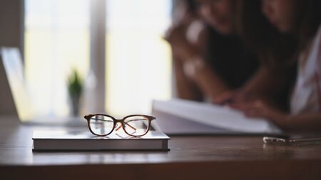 Glasses and book putting together on wooden table with two woman as blurred background....