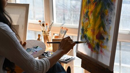 Cropped back image of young beautiful artist holding a brush and drawing on painting canvas while sitting next to the arts accessories putting on small wooden table with studio windows as background.の写真素材