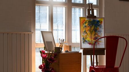 Photo of art studio in morning light. Chair, Wooden table, Painting canvas with easel, Picture frame, Flowers, Vase, Coffee cup, Pencil holder, Paint brush, Windows. Cluttered workplace for artist.の写真素材