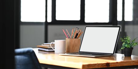 Blank screen computer laptop putting on wooden working desk with pencil holder, stack of books, coffee cup and potted plant over orderly living room as background.の写真素材