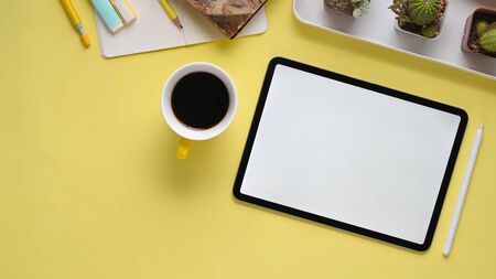 Top view image of colorful workplace. Pencil compass, coffee cup, notebook, marker pens, cactus, stylus pen and computer tablet with white empty screen flat lay on yellow table.の写真素材