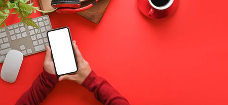 Top view image of hands with red long sleeves holding a empty screen smartphone over orderly working desk. Wireless keyboard, mouse, coffee cup, potted plant, wireless headphone putting on red table.の写真素材