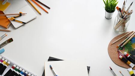 Top view image of painter working desk. Color tube, Potted plant, Paint brush, Stationary, Color palette and Notebook flat lay on white isolated background. Cluttered workspace concept.の写真素材