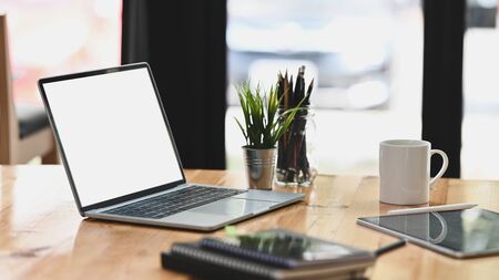 Laptop with white blank screen putting on modern wooden table together with computer tablet, coffee cup, potted plant, notebook. Orderly/Comfortable workplace concept.の写真素材