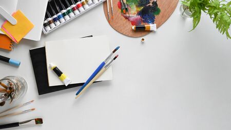 Top view image of painter working desk. Color tube, Potted plant, Paint brush, Stationary, Color palette and Notebook flat lay on white isolated background. Cluttered workspace concept.の写真素材