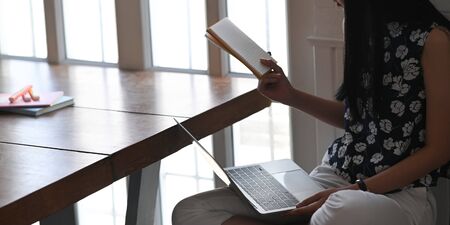 Closeup image of stylish woman holding a book and computer laptop in her hands while sitting at the wooden working desk over comfortable living room as background.の写真素材
