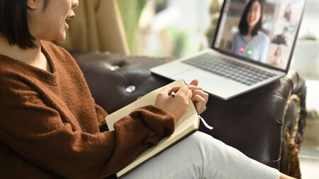 Beautiful woman taking notes while sitting in front her laptop at the leather couch in living room....