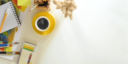 Top view image of ceramic coffee cup putting on center of white working desk and surrounded by painting canvas, paint brush, pencil holder, color tube, note, pen and wild grass. Painter working desk.の写真素材