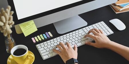Cropped image of hands typing on wireless keyboard that putting on black working desk with computer...