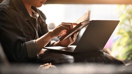 Man holding and reading a book while sitting in front a computer laptop at the leather couch over comfortable living room as background.の写真素材