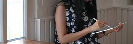 Creative woman holding a computer tablet and stylus pen while sitting at the wooden working desk over comfortable sitting room as background.の写真素材