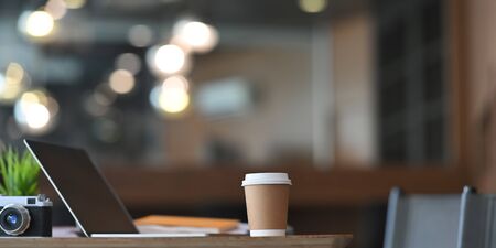 Take-away coffee cup putting on working desk surrounded by computer laptop with black empty screen, camera, potted plant and stack of paperwork over luxury and modern workplace as background.の写真素材