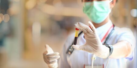 Photo of beautiful female doctor wearing a medical mask and glove showing a blood tube for test while standing at the modern examination room over blurred hospital as background.の写真素材
