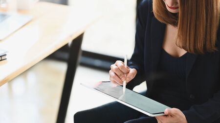 Cropped shot sexy businesswoman holding a computer tablet and stylus pen while sitting at the wooden working desk over comfortable sitting room as background.の写真素材