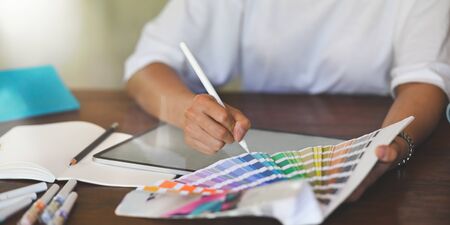 Closeup graphic designer drawing on computer tablet while selecting color from color guide and sitting at wooden working desk that surrounded by office equipment.の写真素材