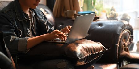 Photo of attractive man in denim shirt working/typing on computer laptop that putting on his lap while sitting at the leather sofa over comfortable living room as background.の写真素材