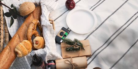 Top view image different sorts of breads in wicker basket that putting on a blanket and surrounded by plastic dish, apple, note, bottle of beer and camera. Picnic equipment on sunny outside concept.の写真素材