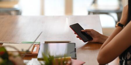 Cropped image of young entrepreneur woman using smartphone while sitting in front her computer tablet that putting on wooden working desk over comfortable sitting room windows as background.の写真素材