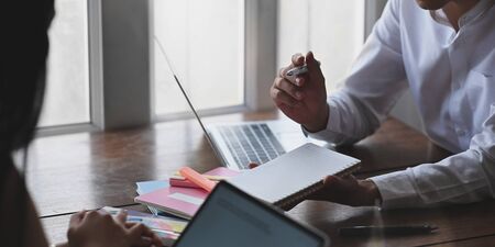 Cropped image of businessman taking notes while sitting in front of computer laptop and his colleague at the wooden working desk over sitting room windows as background.の写真素材